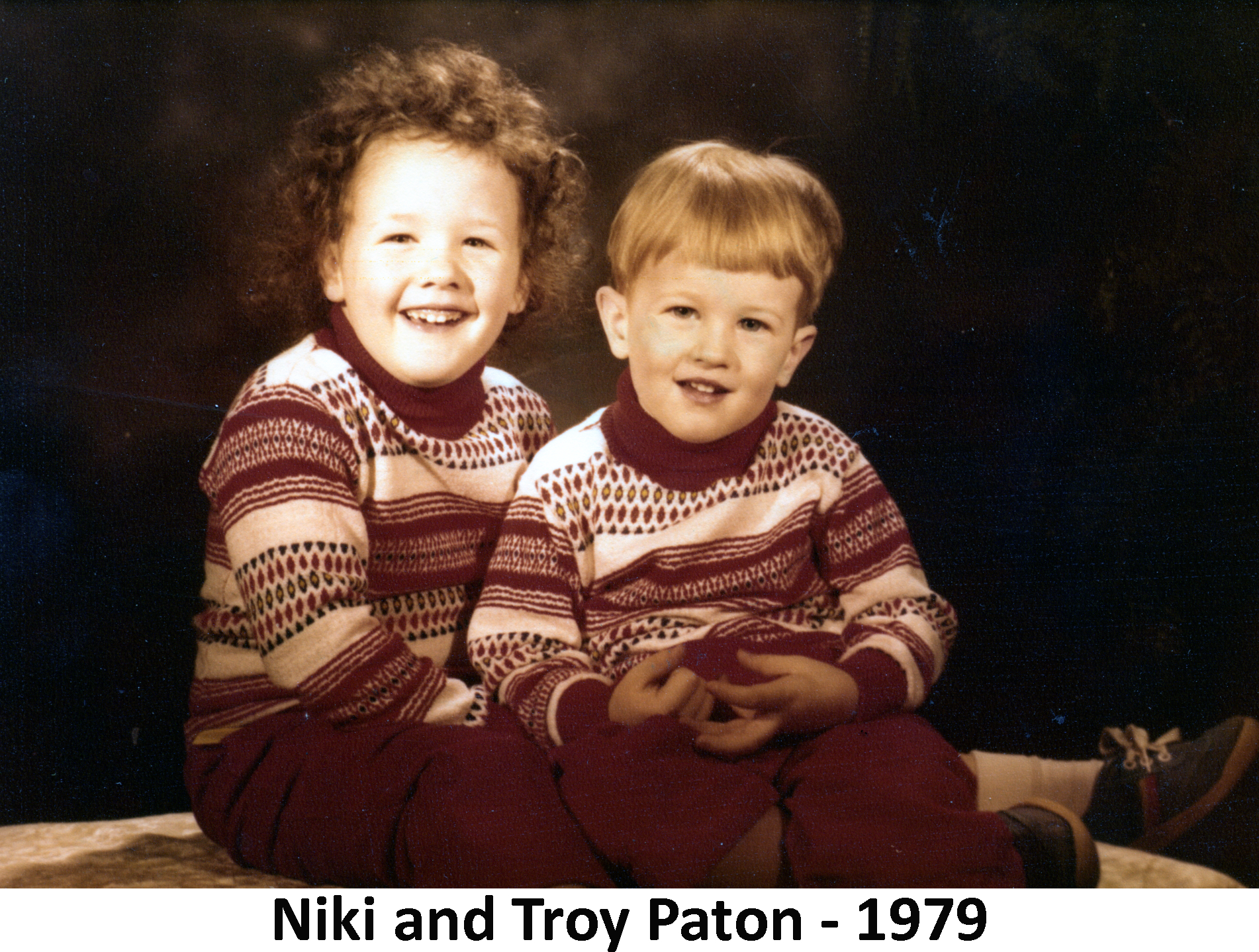 Niki and Troy Paton are sitting together with a dark background in a
              studio photo. Both are wearing matching, red-and-white sweaters.