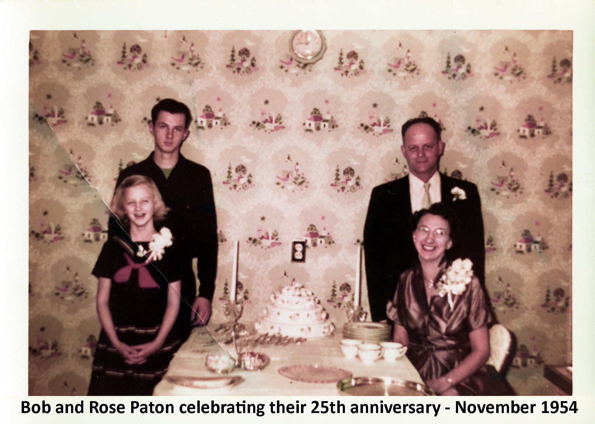 The Paton family is dressed nicely. Gail and Rose are seated at a table
          with a fancy cake on it and Guy and Robert are standing in behind them.