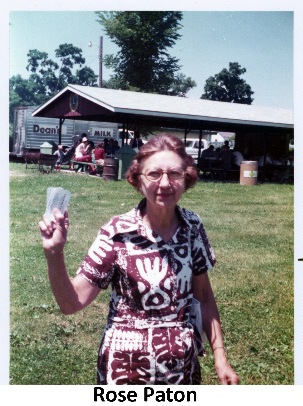 Rose Paton standing in Veterans Park and holding several pieces of paper, 
               possibly raffle tickets