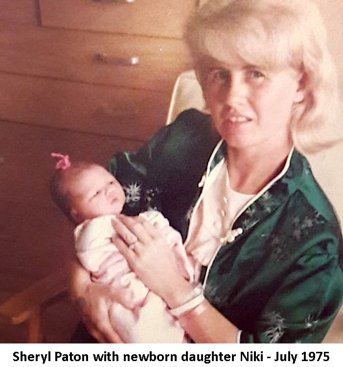 Sheryl Paton is holding her newborn baby Niki. 
             Sheryl is wearing a green robe and Niki has a pink ribbon in her hair.
