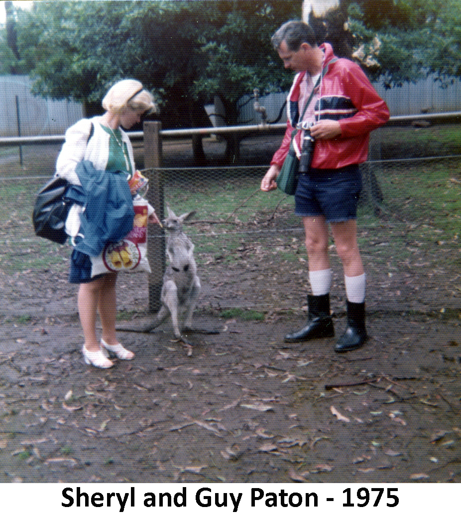 Sheryl and Guy Paton feeding treats to a wallaby, that stands
         about thigh-high between them. There’s a fence and a tree behind them.