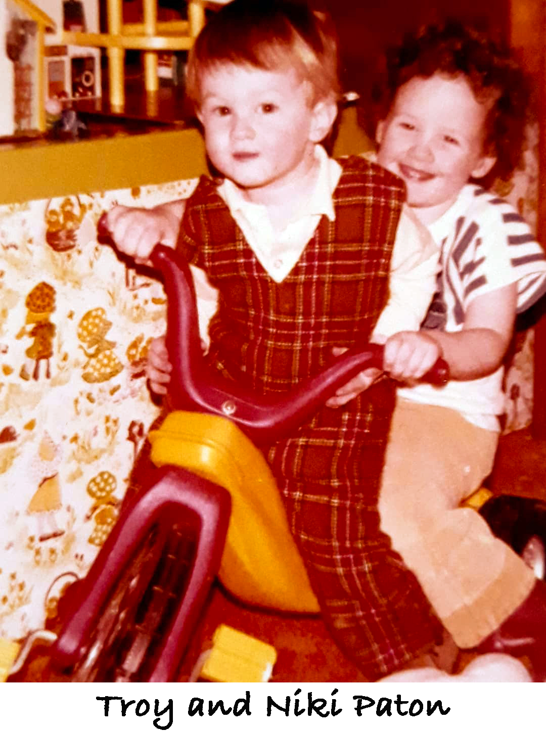 Troy and Niki Paton are riding on a “Big Wheel” tricycle.
               Troy is wearing a bright red coverall.