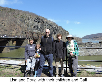 The family is standing in front of the river on a sunny day. 
                 People are walking across the footbridge on the left