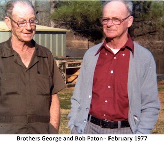 George and Robert Paton standing in a yard with a large utility shed and pine trees