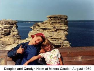 Douglas and Carolyn Holm standing at a wall                  with rock tower and Lake Superior in the background