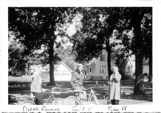 The three girls are looking toward the camera.                They have the old-fashioned, balloon-tire bikes. 