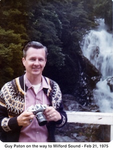 Guy is wearing a sweater and holding a camera in front of green 
              bushes and a waterfall