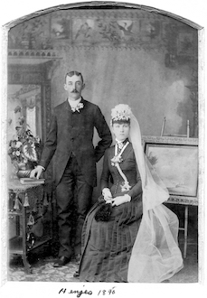Fred Henjes and Ella Duntemann studio photo. She’s sitting and wearing a dark dress with a white train. He’s standing with his hand on a book