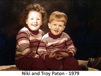 Niki and Troy Paton are sitting together with a dark background in a              studio photo. Both are wearing matching, red-and-white sweaters.