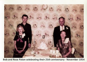 The Paton family is dressed nicely. Gail and Rose are seated at a table          with a fancy cake on it and Guy and Robert are standing in behind them.