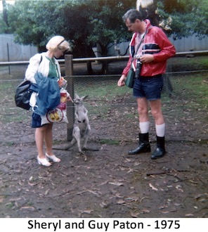 Sheryl and Guy Paton feeding treats to a wallaby, that stands
      about thigh-high between them. There’s a fence and a tree behind them.