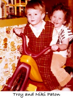 Troy and Niki Paton are riding on a “Big Wheel” tricycle.
               Troy is wearing a bright red coverall. 