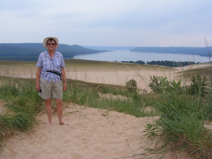 Gail faces the camera. In the distance behind
               her is Glen Lake. There is sand at the top, but 
               surrounded by dune vegetation. A wide bench of sand
               is behind and below her. 