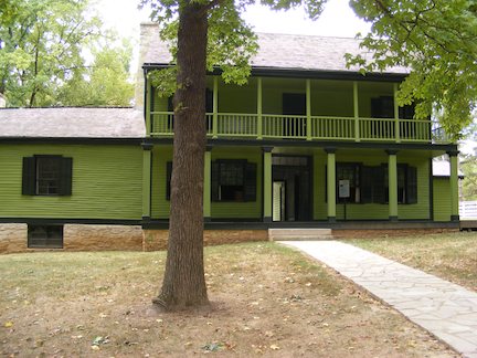The house is
              painted green. The main part is two stories
              with a covered porch and a covered balcony 
              above it. To the left is a one-story wing
              with no porch.