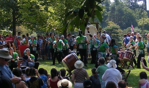Many drummers in green shirts
      are surrounded by an audience