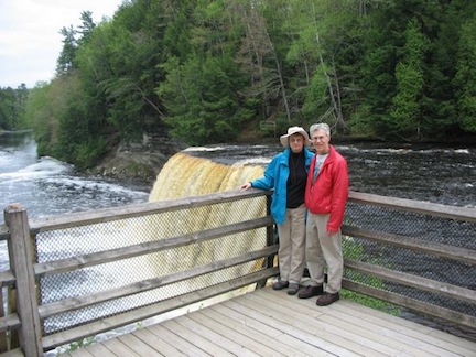 Gail 
           and Al stand on a platform overlooking 
           the upper falls at Tahquamenon. The root-beer
           colored water pours over the 50-foot drop
           behind them