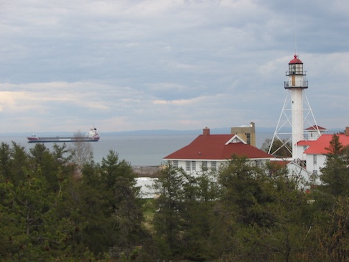 The         freighter is sailing to the left. The light sits atop         a large, white cylinder on the right of the photo.          Red-roofed, two-story, white buildings are on both         sides of the light tower. Trees are seen at the bottom         of the photo.