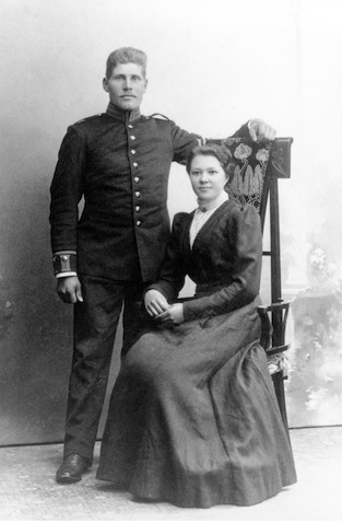 Victor Holm is standing to the left of  
           Frida, who is sitting in a chair with a tall, straight back. 
           He is wearing his Swedish army uniform and she is wearing a 
           long, dark dress. There is nothing else in the photo.